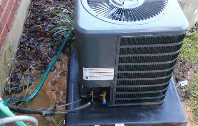 An outdoor air conditioning condenser unit installed on a concrete pad next to a house, with visible refrigerant lines connected to its base.