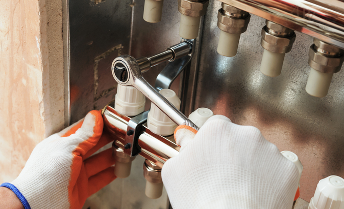 The image shows a technician using a wrench to adjust or repair a manifold in a hydronic heating system, wearing protective gloves for safety.